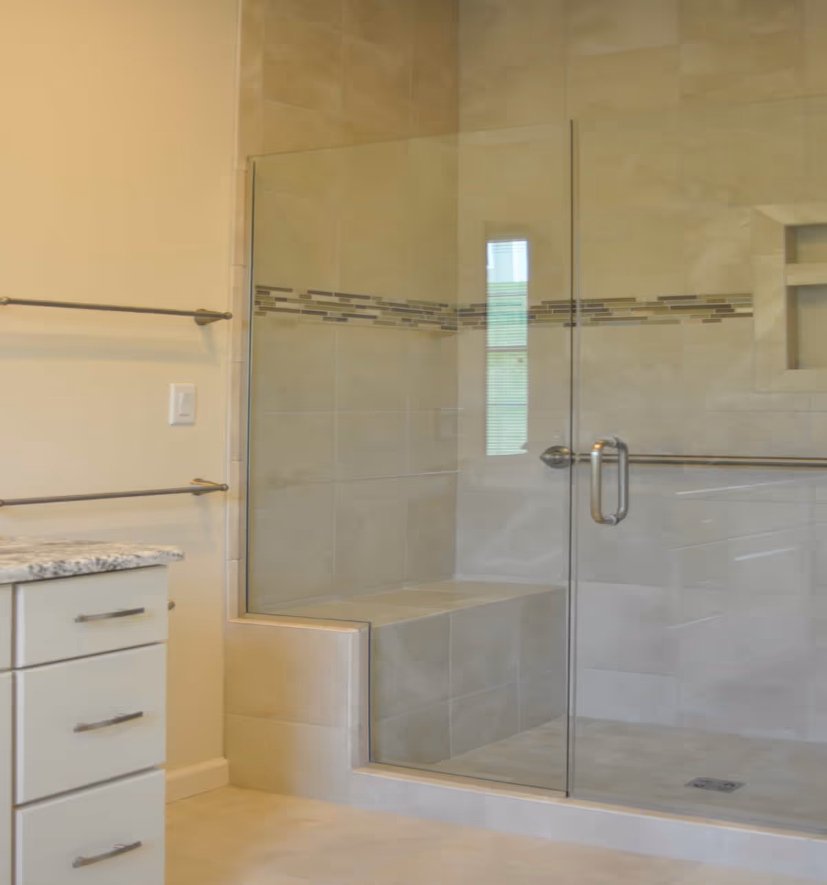 A modern bathroom featuring a glass-enclosed shower with a built-in bench and decorative tile accent strip. The shower has a metal handle and a towel bar on the outside. To the left, there is a countertop with drawers and two towel bars mounted on the wall.