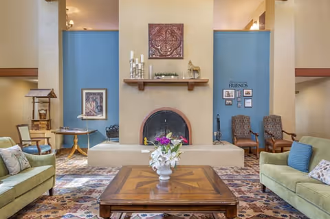 Comfortable communal living room with sofas and armchairs arranged around a wooden coffee table topped with flowers and a central fireplace set against blue accent walls.