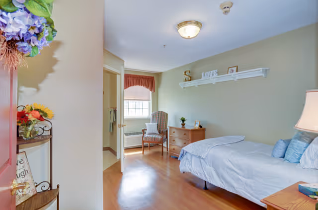 A cozy bedroom in a senior living facility featuring a single bed with light blue bedding and pillows, a wooden nightstand with a lamp, a wooden dresser with decorative items, a comfortable armchair near a window with a red valance, and a shelf on the wall with decorative letters spelling 'LOVE'. The room has light green walls and wooden flooring.