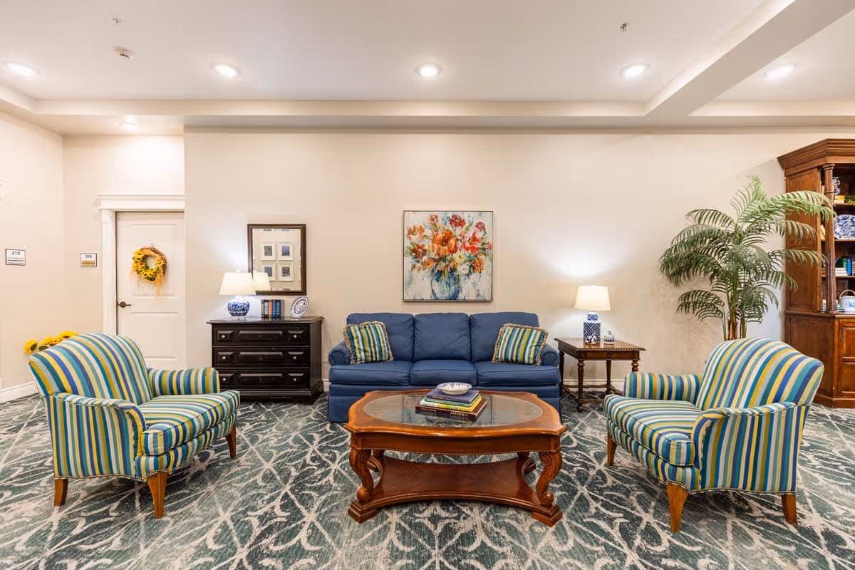 A cozy living room area in an assisted living facility featuring a blue sofa with two striped cushions, two matching striped armchairs, a wooden coffee table with books and a bowl, two side tables with blue and white lamps, a dark wooden chest of drawers with decorative items, a large potted plant, and a colorful floral painting on the wall.