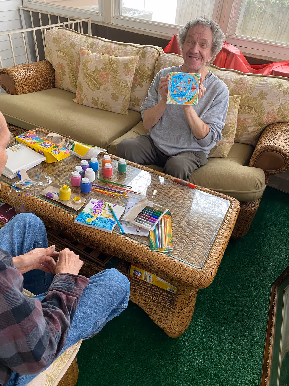 A person seated on a sofa holding a painted tile and smiling while art supplies cover a wicker coffee table.