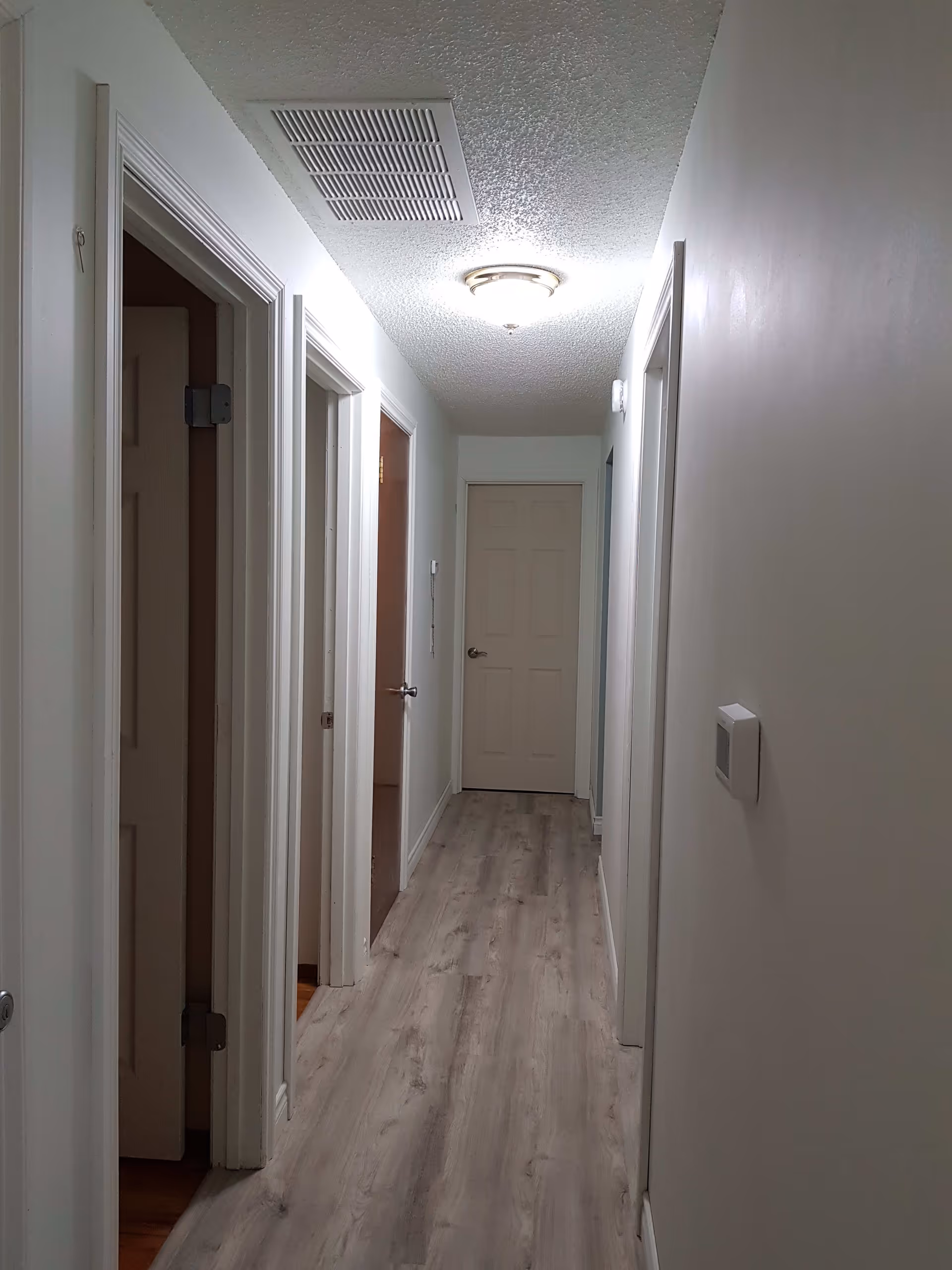 A narrow hallway with light-colored walls and wood-patterned flooring. There are several doorways on both sides of the hallway, and a closed door at the end. A ceiling light fixture illuminates the space.