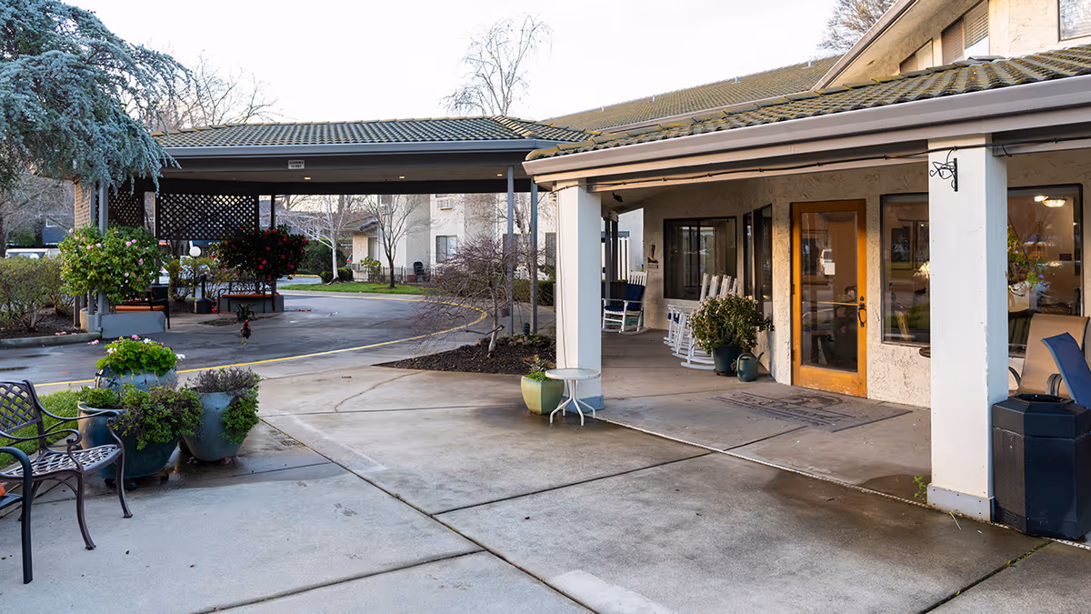 Outdoor view of the entrance area of Provincial Chico Senior Living facility featuring a covered driveway, potted plants, benches, rocking chairs on the porch, and a wooden door with windows.