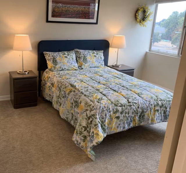 A bedroom with a bed covered in a floral patterned comforter and matching pillows. The bed has a dark upholstered headboard and is flanked by two dark wooden nightstands, each with a lit table lamp. A framed picture hangs on the wall above the bed, and a window with a wreath on the wall next to it lets in natural light.