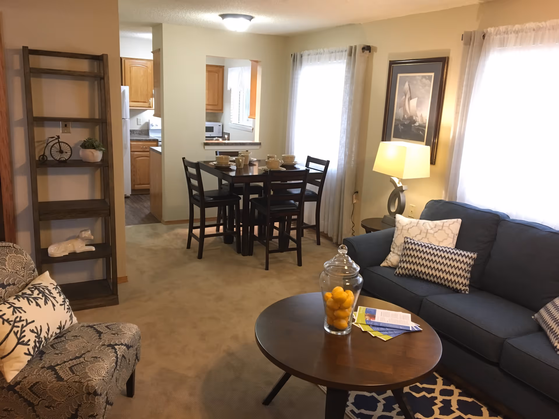 A cozy living and dining area in a senior living facility. The living room features a blue sofa with patterned pillows, a round wooden coffee table with a glass jar filled with lemons and some brochures, and a patterned armchair with a decorative pillow. A wooden shelving unit with decorative items is against the wall. The dining area has a dark wooden table set with four chairs and tableware. The kitchen is visible through a pass-through window and an open doorway, showing wooden cabinets and white appliances. Light beige walls and carpet, with windows covered by sheer curtains, brighten the space.