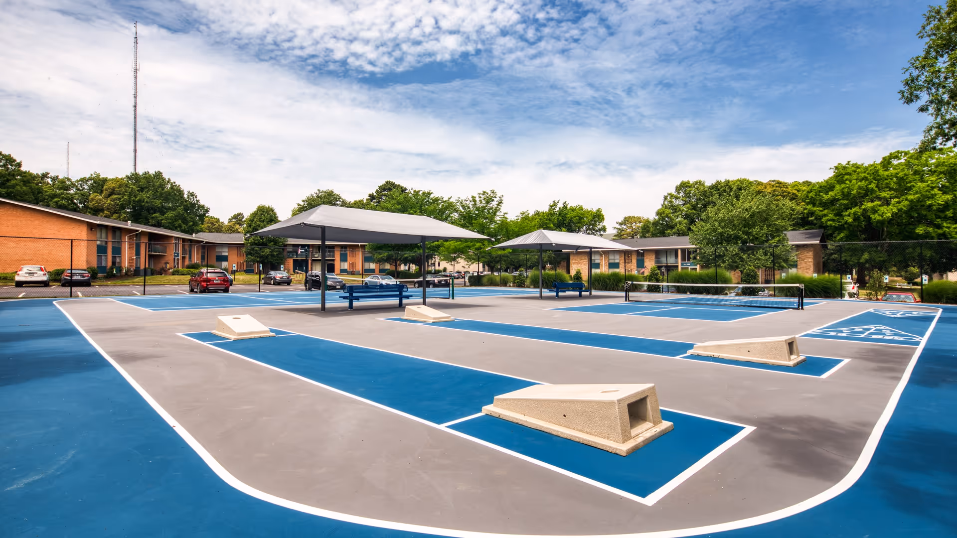 Outdoor shuffleboard courts with blue and gray playing surfaces, shaded seating areas with benches, surrounded by trees and a parking lot with cars. Brick apartment buildings are visible in the background under a partly cloudy sky.