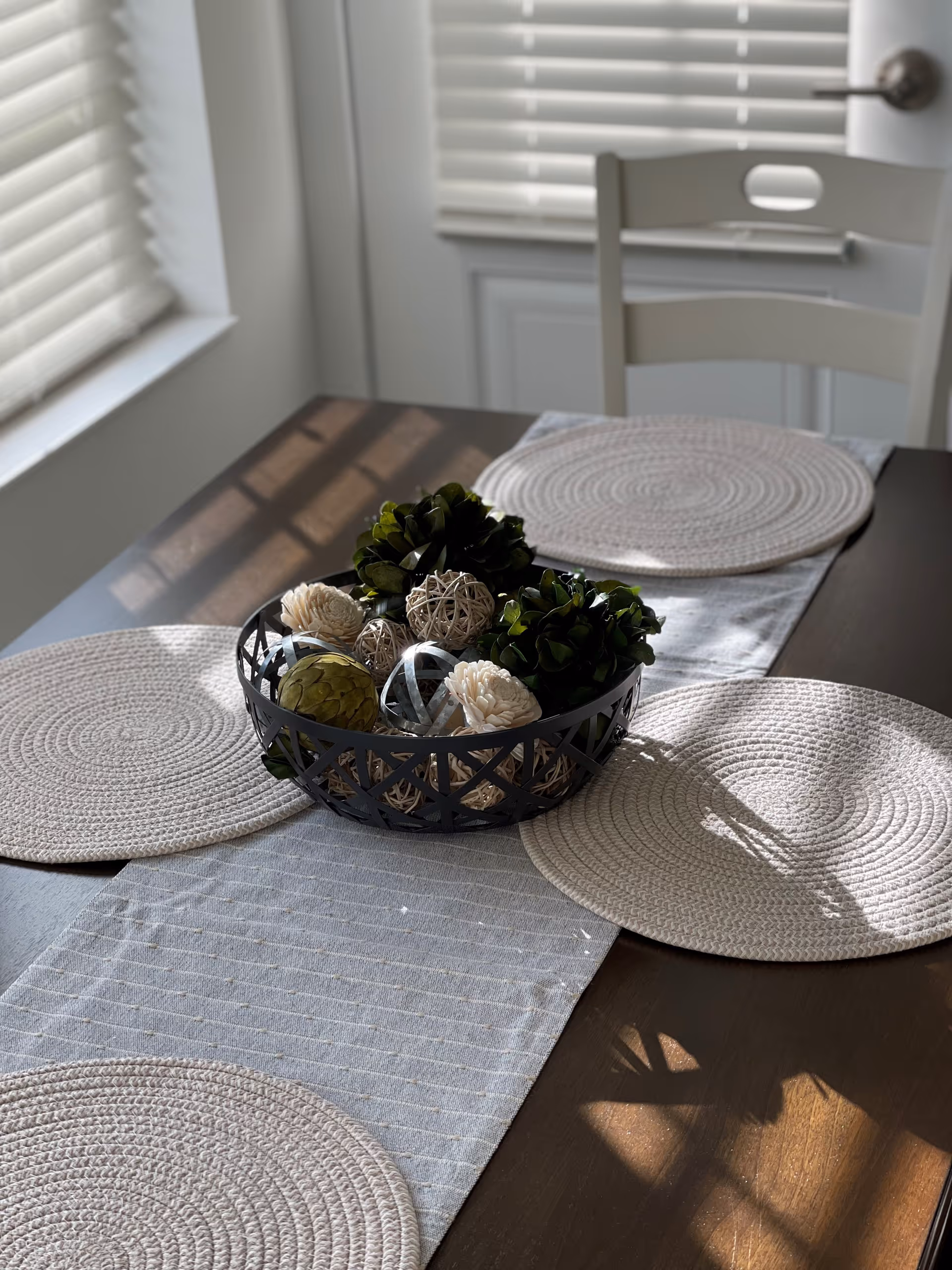 A wooden dining table set with four round woven placemats and a gray table runner. In the center of the table is a decorative black basket filled with assorted decorative balls made of natural materials. Sunlight streams through nearby window blinds, casting shadows on the table.