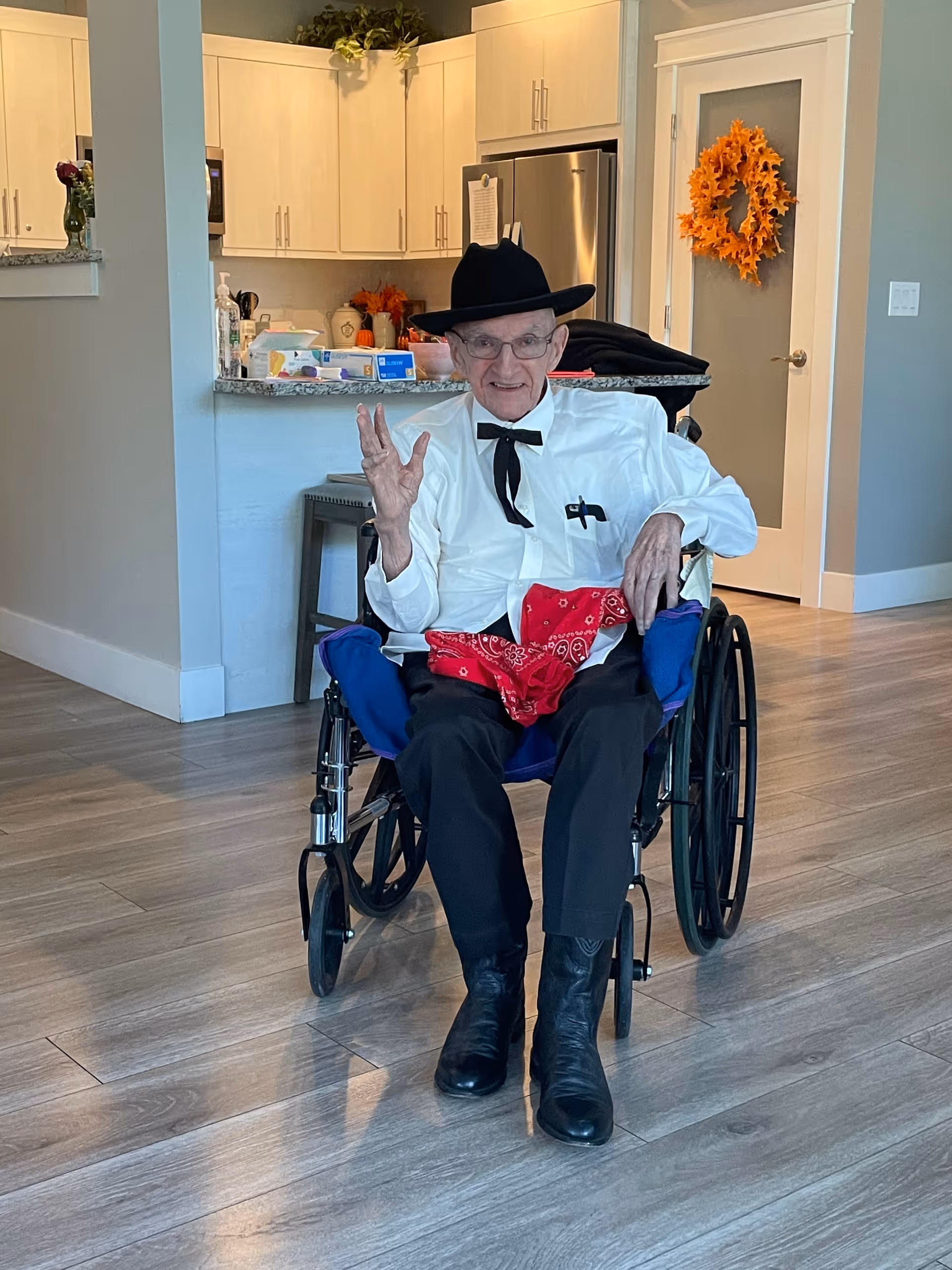 An elderly man in a wheelchair wearing a black cowboy hat and bow tie sits in a modern open-plan living area with a kitchen in the background.