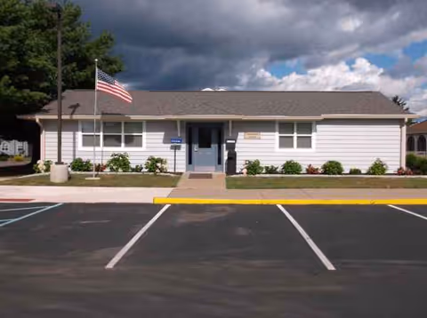 Front view of a single-story building with American flag, parking lot, and landscaped shrubs.