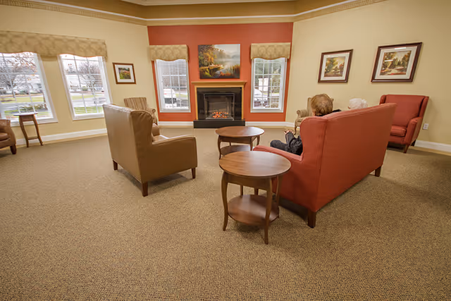 Communal living room with sofas, armchairs, side tables and a fireplace against a red accent wall with windows.