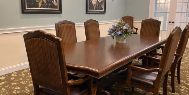 Long wooden dining table with eight high-backed chairs and a floral centerpiece in a formal dining room.