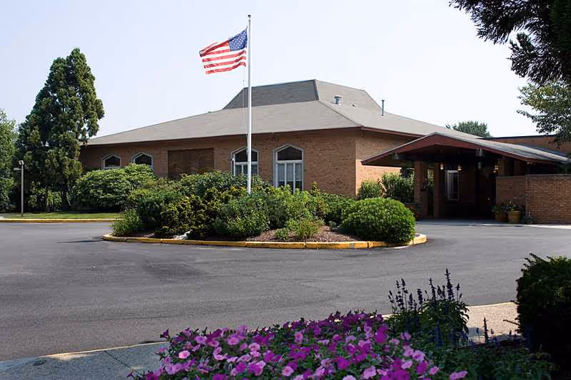 Exterior view of a brick building with a gray roof, an American flag on a flagpole in front, surrounded by green bushes and trees, with a flower bed of purple flowers in the foreground.