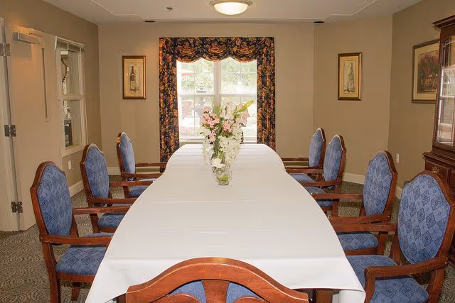 A dining room with a long rectangular table covered with a white tablecloth. There are eight wooden chairs with blue upholstered seats and backs arranged around the table. A vase with pink and white flowers is placed in the center of the table. The room has beige walls, a window with floral curtains, and framed artwork on the walls.