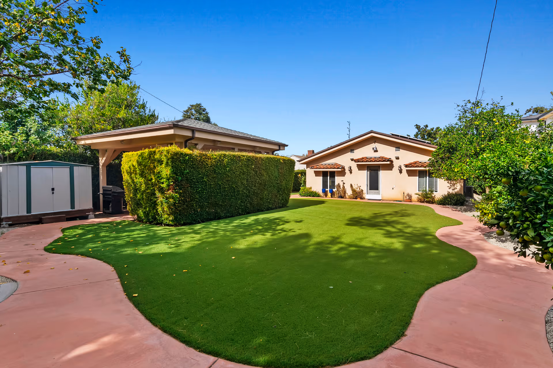 Outdoor area of a senior living facility with a well-maintained green lawn, a curved pinkish concrete pathway, a small shed, a covered patio area, and a single-story building with beige walls and red-tiled awnings under a clear blue sky.