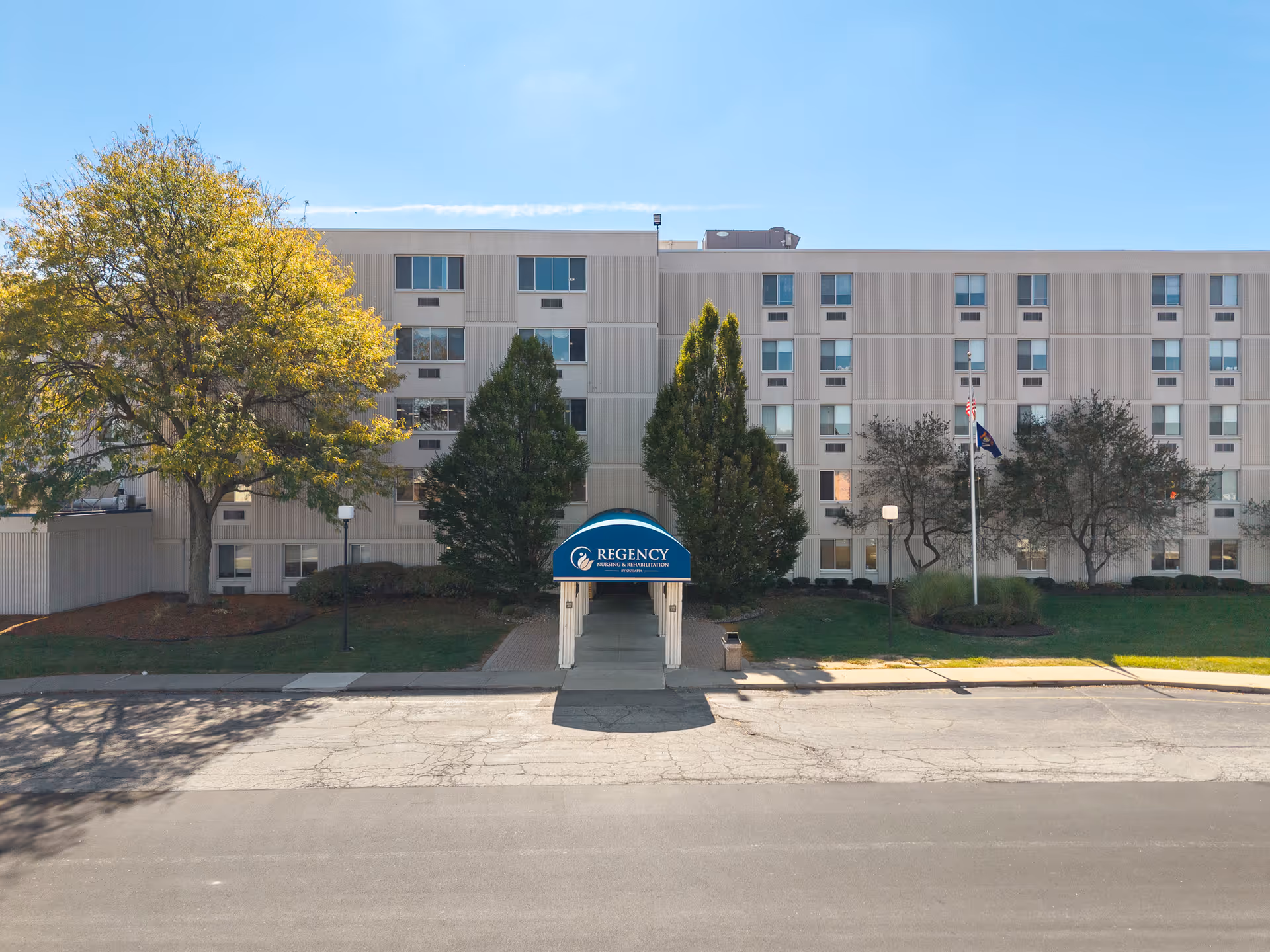 Front exterior of a multi-story senior living building with a blue 'Regency' entrance canopy, trees, and flagpoles.