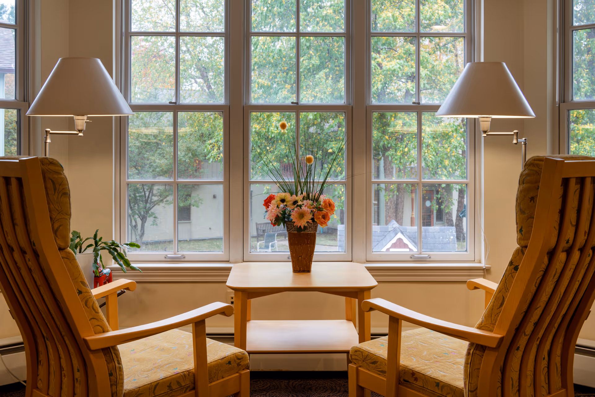 Two wooden armchairs with patterned cushions facing each other with a small wooden table between them, holding a vase of colorful flowers. Behind the table is a large window with multiple panes showing green trees outside. Two lamps with white shades are positioned behind each chair.
