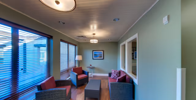 A cozy living room area with green walls, wooden flooring, and a white ceiling. The room features two armchairs and a loveseat with red cushions, a small glass side table with a lamp, a rectangular ottoman, and a framed picture on the far wall. Large windows with blinds allow natural light to enter the space.