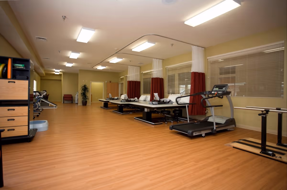 A spacious rehabilitation or physical therapy room with wooden flooring, several treatment tables separated by curtains, a treadmill, parallel bars for walking exercises, and storage cabinets with exercise equipment. The room is well-lit with ceiling lights and has large windows with blinds.