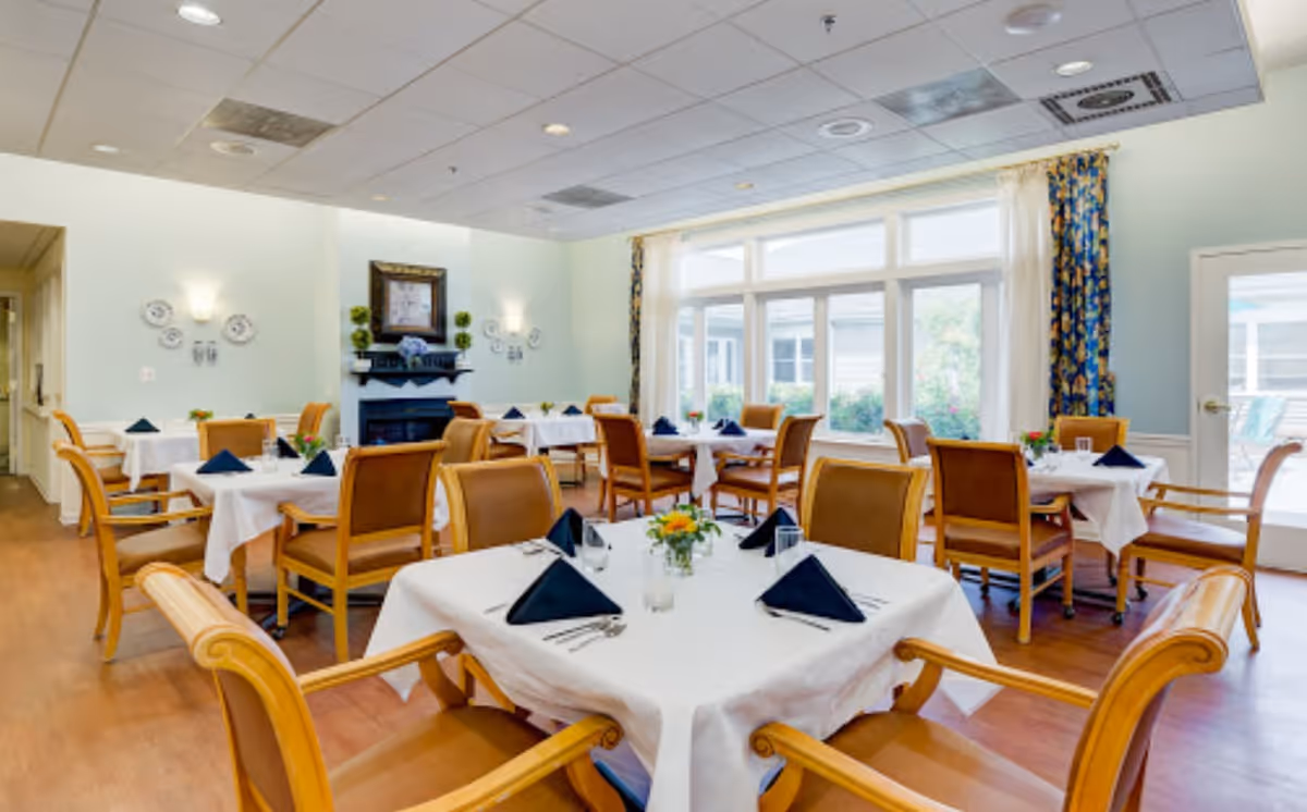 A bright dining room with several tables covered in white tablecloths, each set with navy blue folded napkins, silverware, and small flower arrangements. The room has wooden chairs with armrests, large windows with patterned curtains letting in natural light, and a decorative fireplace with a framed picture above it.