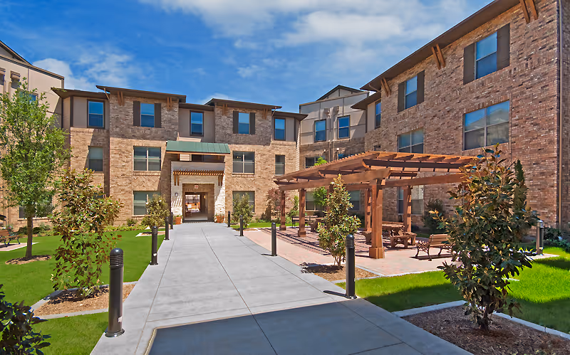 Outdoor courtyard area of a senior living facility with a paved walkway leading to a brick building entrance. The courtyard features green grass, small trees, and a wooden pergola with benches and tables underneath. The sky is clear and blue.