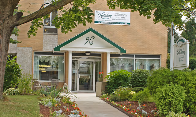Entrance to Holiday Retirement senior independent living and retirement community building with a glass door under a small green and white awning. There are well-maintained flower beds and shrubs along the walkway leading to the entrance. A sign with the community name and contact information is visible above the door and another sign is on the right side near the bushes.