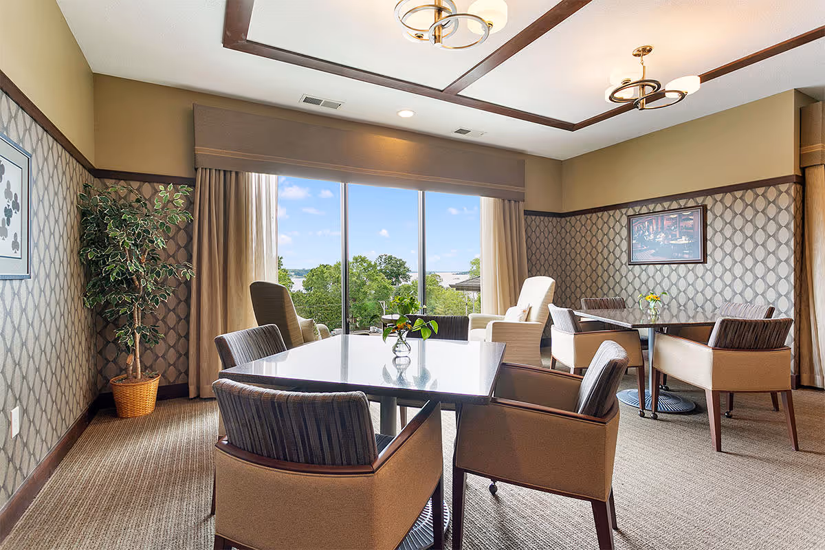 A well-lit dining room with two tables, each surrounded by four cushioned chairs. The room features patterned wallpaper, a large window with a view of trees and a blue sky, beige carpeted floor, and two ceiling light fixtures. There is a potted plant in the corner and framed artwork on the walls.