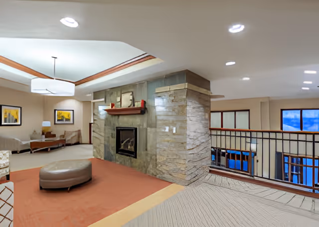 A spacious living area featuring a stone fireplace with a wooden mantel, a round ottoman on a red carpet, beige walls with framed artwork, and a seating area with chairs and a table. The ceiling has recessed lighting and a modern hanging light fixture. There is a railing overlooking a lower level with large windows showing a blue sky outside.