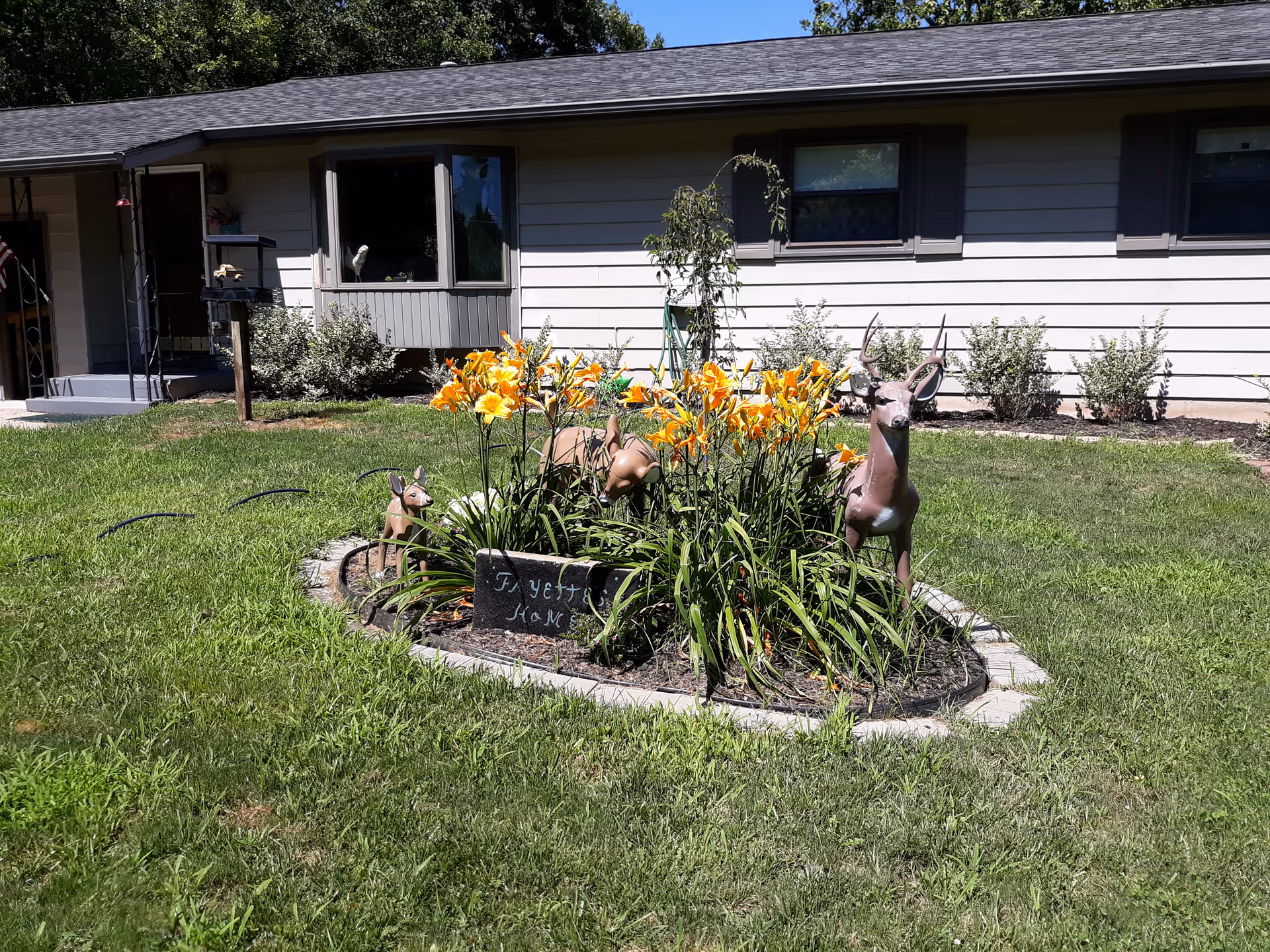 A garden area in front of a single-story building with beige siding and dark shutters. The garden features bright orange flowers and three decorative deer statues. A small sign in the garden reads 'Fayette's Home'. The surrounding lawn is green and well-maintained.