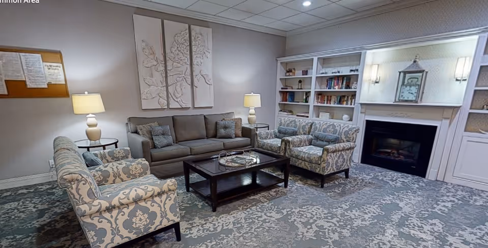 A cozy common area with a gray sofa, two patterned armchairs, a dark wooden coffee table, two side tables with lamps, a fireplace with a clock above it, and built-in bookshelves filled with books and decorative items. The room has a patterned carpet and soft lighting.