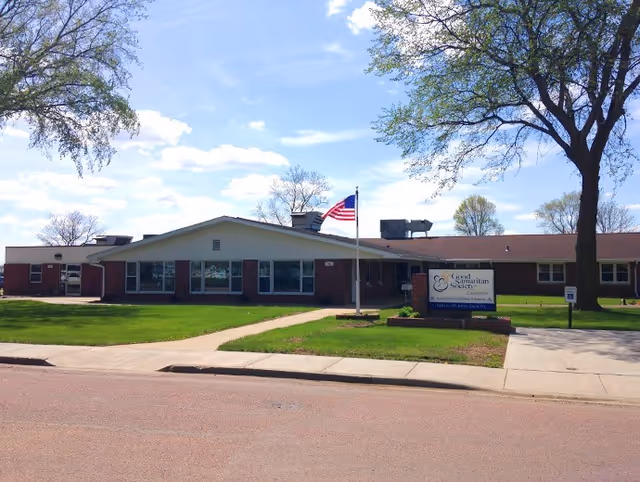 Single-story brick building with a sloped roof, an American flag on a flagpole in front, and a sign that reads Good Samaritan Society - Canistota. The building is surrounded by green grass, trees, and a clear blue sky with some clouds.