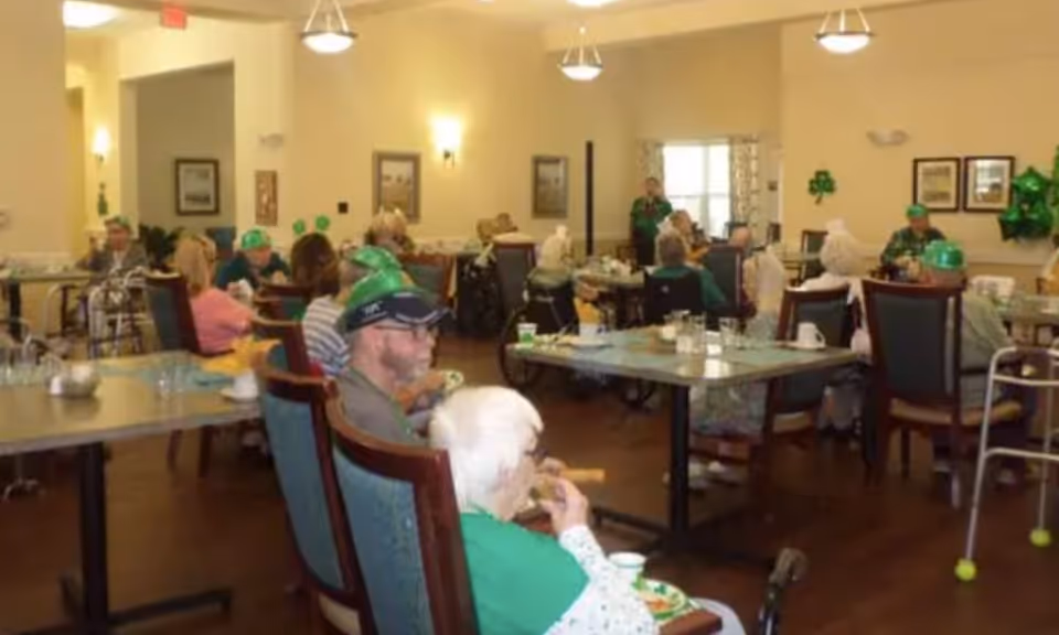 A dining room in an assisted living facility with elderly residents seated at tables. Some residents are wearing green hats and festive decorations are visible on the walls, suggesting a celebration or event. The room has warm lighting and framed pictures on the walls.