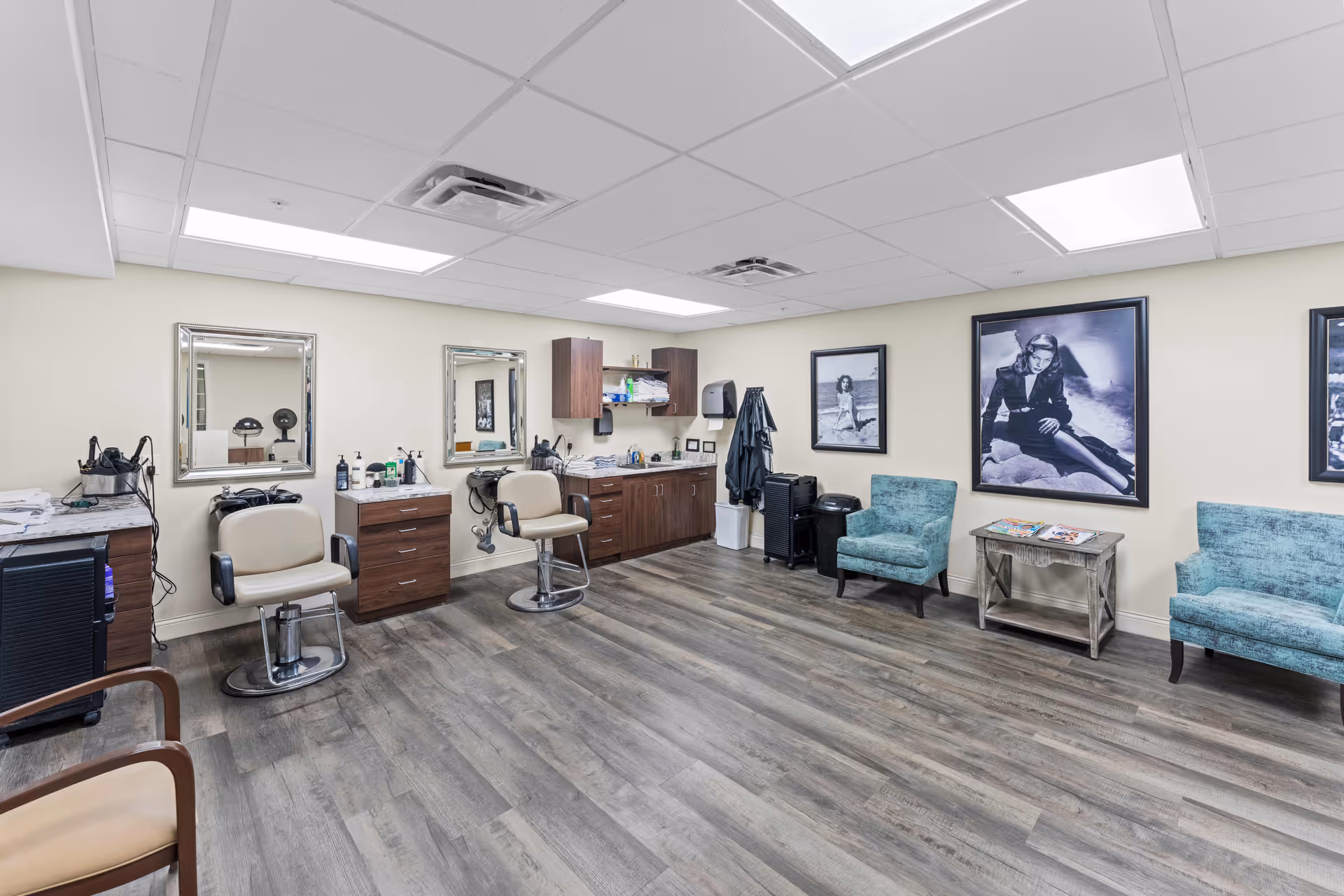 A spacious salon area in a senior living facility with two salon chairs in front of mirrors, wooden cabinets and countertops with hair care products, and two teal upholstered chairs with a small table holding magazines. Black and white framed photographs decorate the walls.