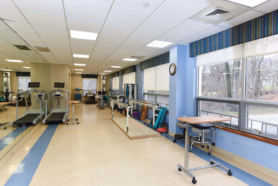 A rehabilitation room with exercise equipment including treadmills, parallel bars for walking practice, and various therapy tools. The room has large windows with blinds, blue walls, a clock on the wall, and a mirrored wall reflecting the equipment.