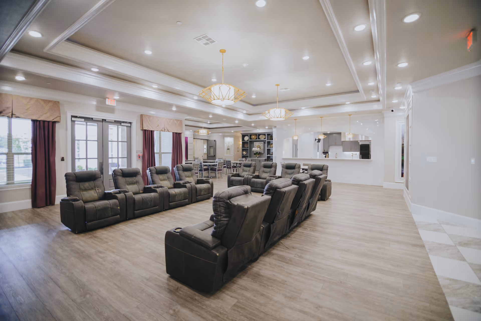 A spacious, well-lit common area with two rows of dark reclining chairs facing each other. The room features wood flooring, large windows with curtains, and modern ceiling lights. In the background, there is a dining area with tables and chairs, and a kitchen area with a counter and pendant lights.