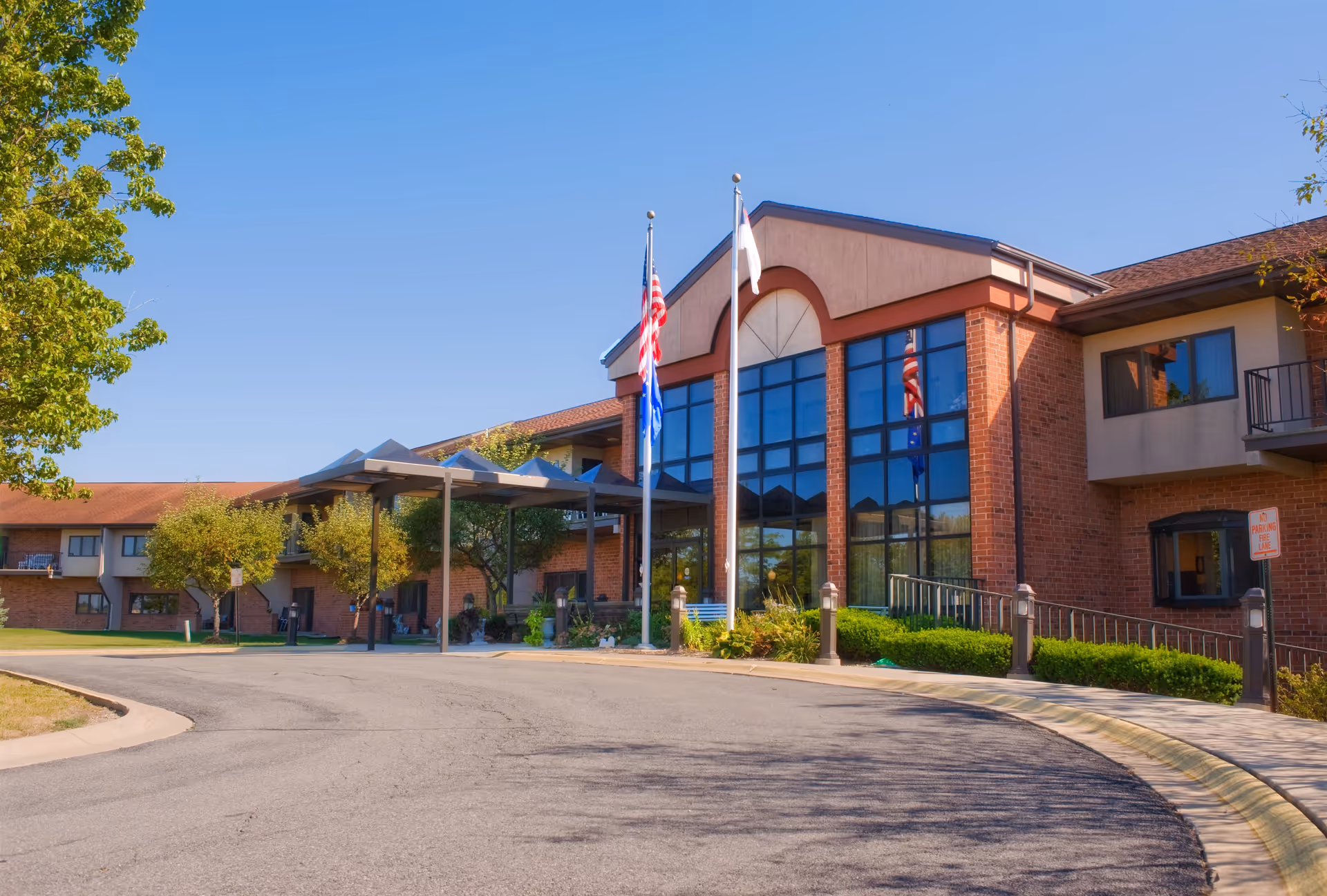 Exterior view of Crown Point Christian Village building with large windows, brick walls, and a covered entrance. Three flagpoles with flags are positioned in front of the building, surrounded by trees and bushes under a clear blue sky.