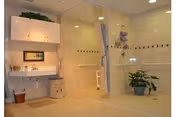 A spacious, accessible bathroom with a wall-mounted sink and mirror on the left, a shower area with a curtain and grab bars on the right, beige tiled walls and floor, and two potted plants placed on the floor.