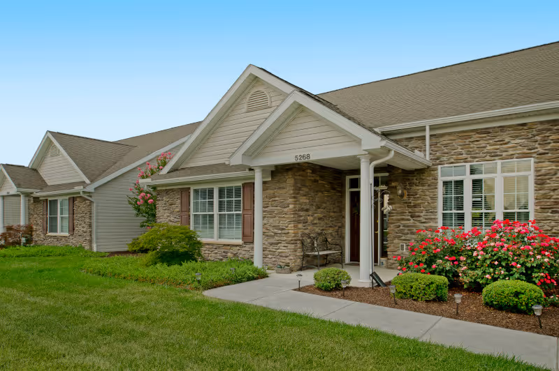 Front exterior of a single-story senior living residence with a stone facade, covered porch, address number 5268, and landscaped lawn and flowerbeds.