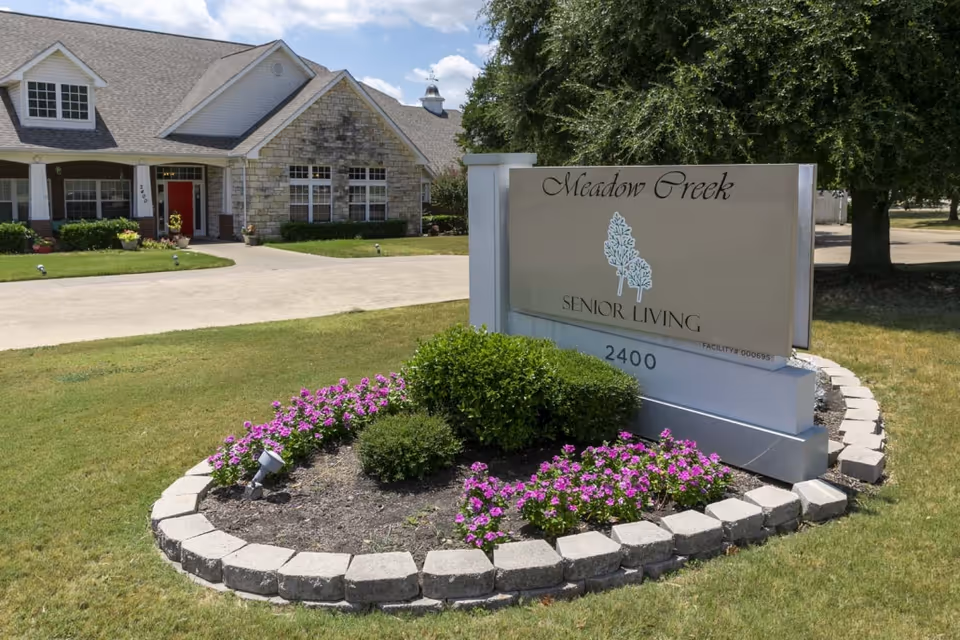 Exterior view of Meadow Creek Senior Living facility showing a large sign with the facility name and address 2400, surrounded by a flower bed with purple flowers and green shrubs. In the background, there is a building with a stone and brick facade, a red front door, and a driveway.