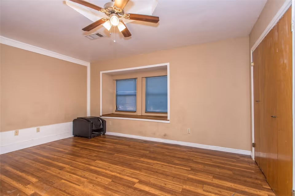 Empty room with hardwood floors, a ceiling fan, a recessed window seat with blinds, and a closed wooden closet door.