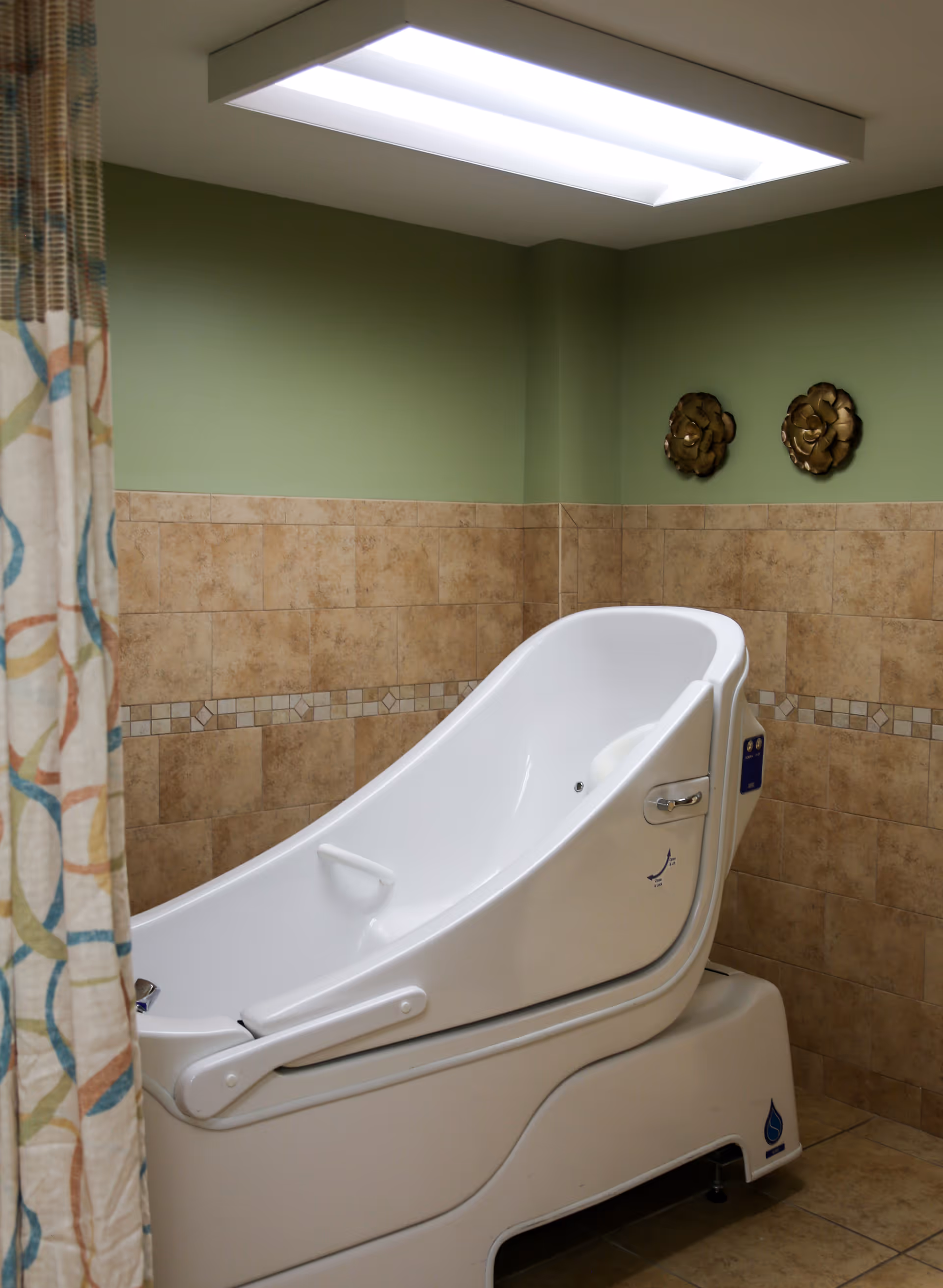 A modern white assisted bathing tub in a tiled room with green walls and a patterned shower curtain partially visible on the left side. The tub is designed for easy access and safety, with handles and controls on the side. Two decorative wall hangings are mounted on the green wall above the tub.