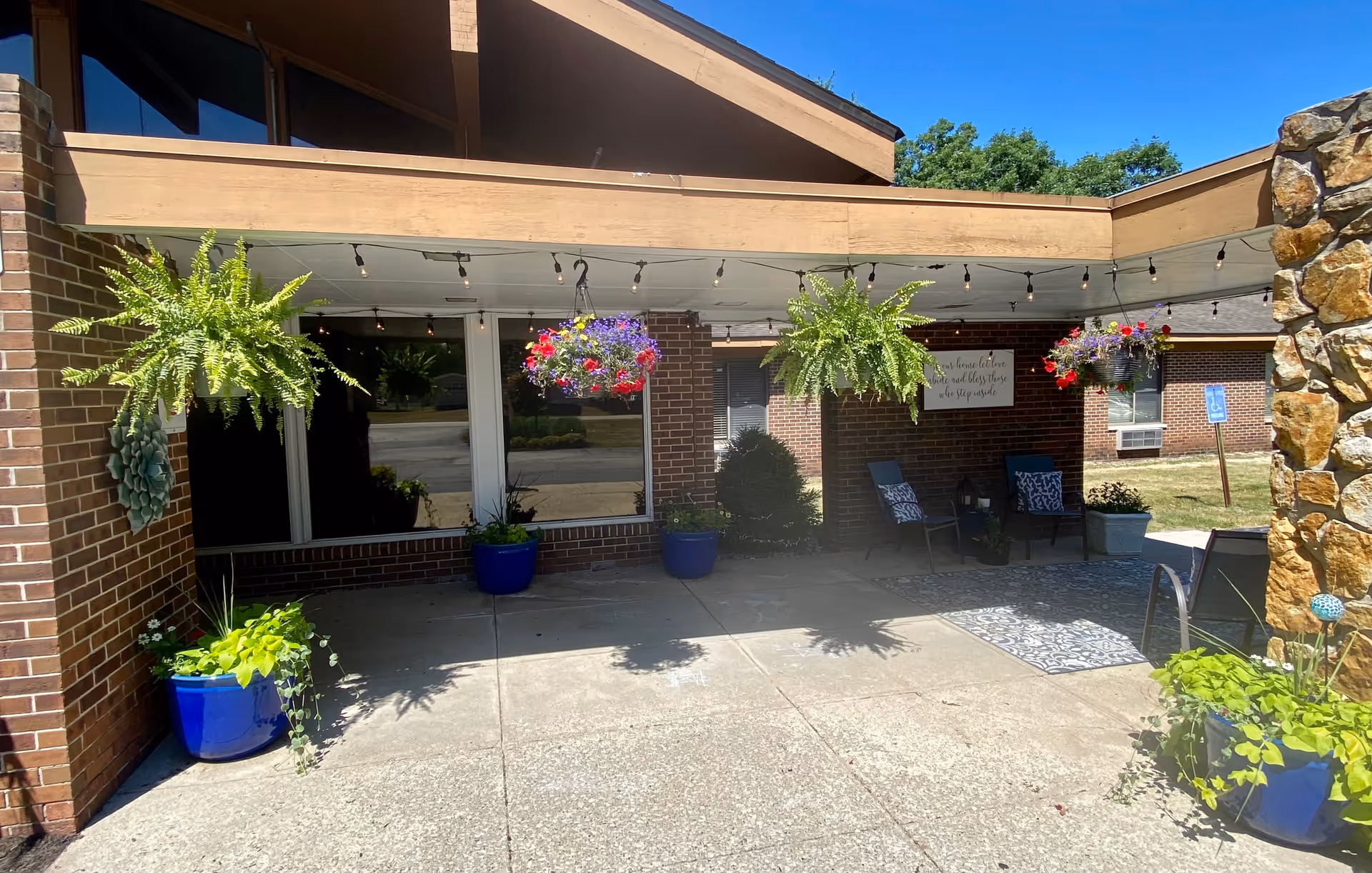 Outdoor covered patio area with hanging flower baskets and potted plants. There are two chairs with cushions and a small table on a patterned outdoor rug. The building has brick walls and a stone pillar, with string lights hanging from the ceiling.