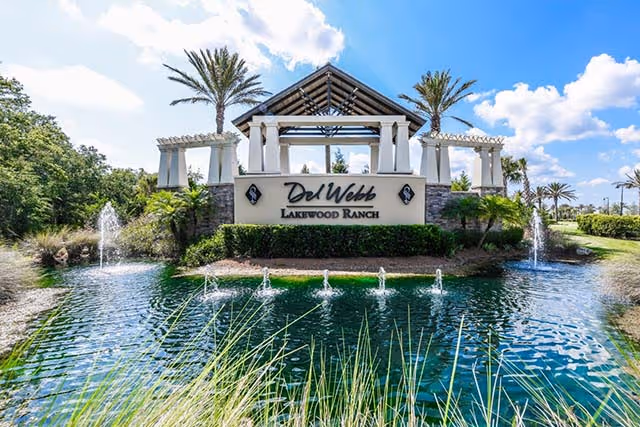 Entrance sign reading 'Del Webb Lakewood Ranch' with a pavilion, palm trees, and fountains over a pond.