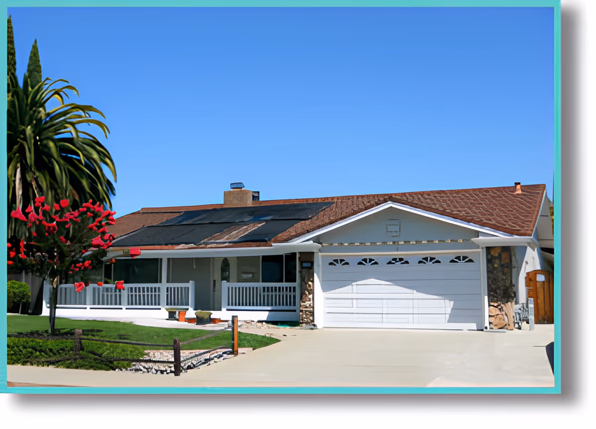 Front exterior view of a single-story residential building with a white garage door, a porch with white railings, a driveway, green lawn, and a tree with red flowers under a clear blue sky.