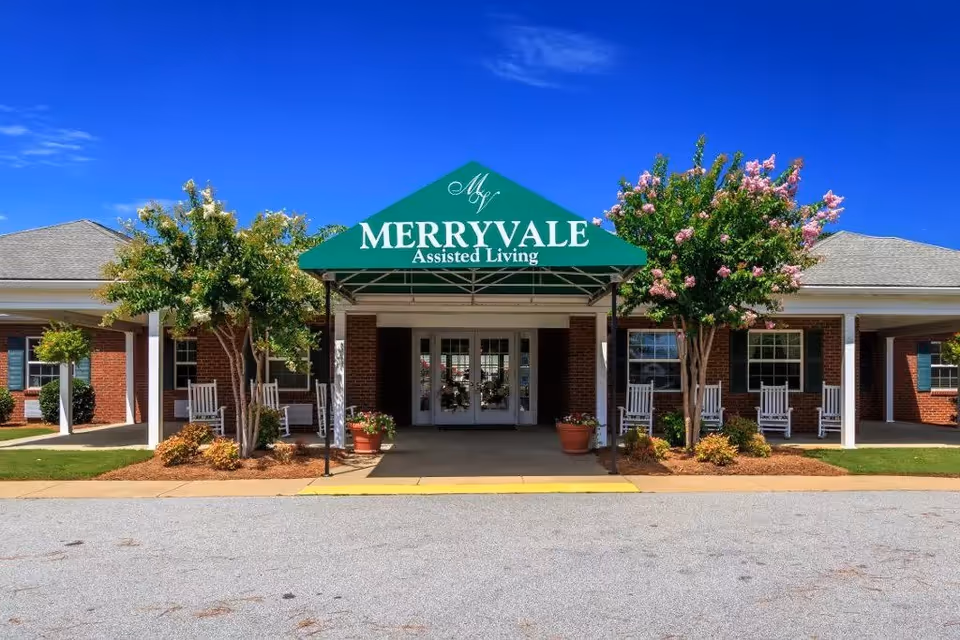 Front entrance of Merryvale Assisted Living facility with a green canopy displaying the facility name. The building is brick with white trim, and there are flowering trees and rocking chairs on the porch under a clear blue sky.