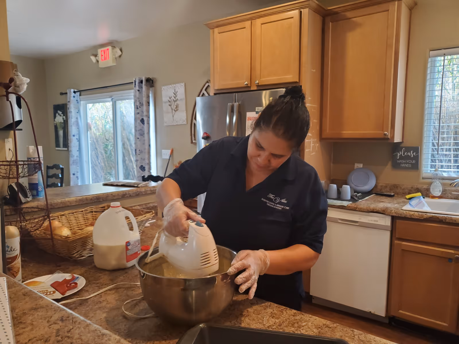 A woman wearing gloves uses an electric hand mixer to mix ingredients in a large metal bowl in a kitchen. The kitchen has wooden cabinets, a stainless steel refrigerator, a dishwasher, and a window with blinds. A gallon of milk and a basket with bread are on the counter nearby.