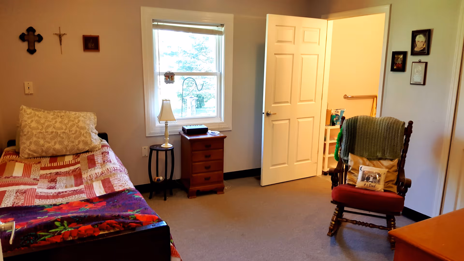 A cozy bedroom with a single bed covered in a patchwork quilt and a large pillow. Next to the bed is a small round table with a lamp and a wooden nightstand with drawers. A window with a view of greenery outside is centered on the wall. An open door leads to another room, possibly a bathroom. A wooden rocking chair with a green knitted blanket and a pillow sits near the door. Several framed pictures and religious crosses hang on the walls.