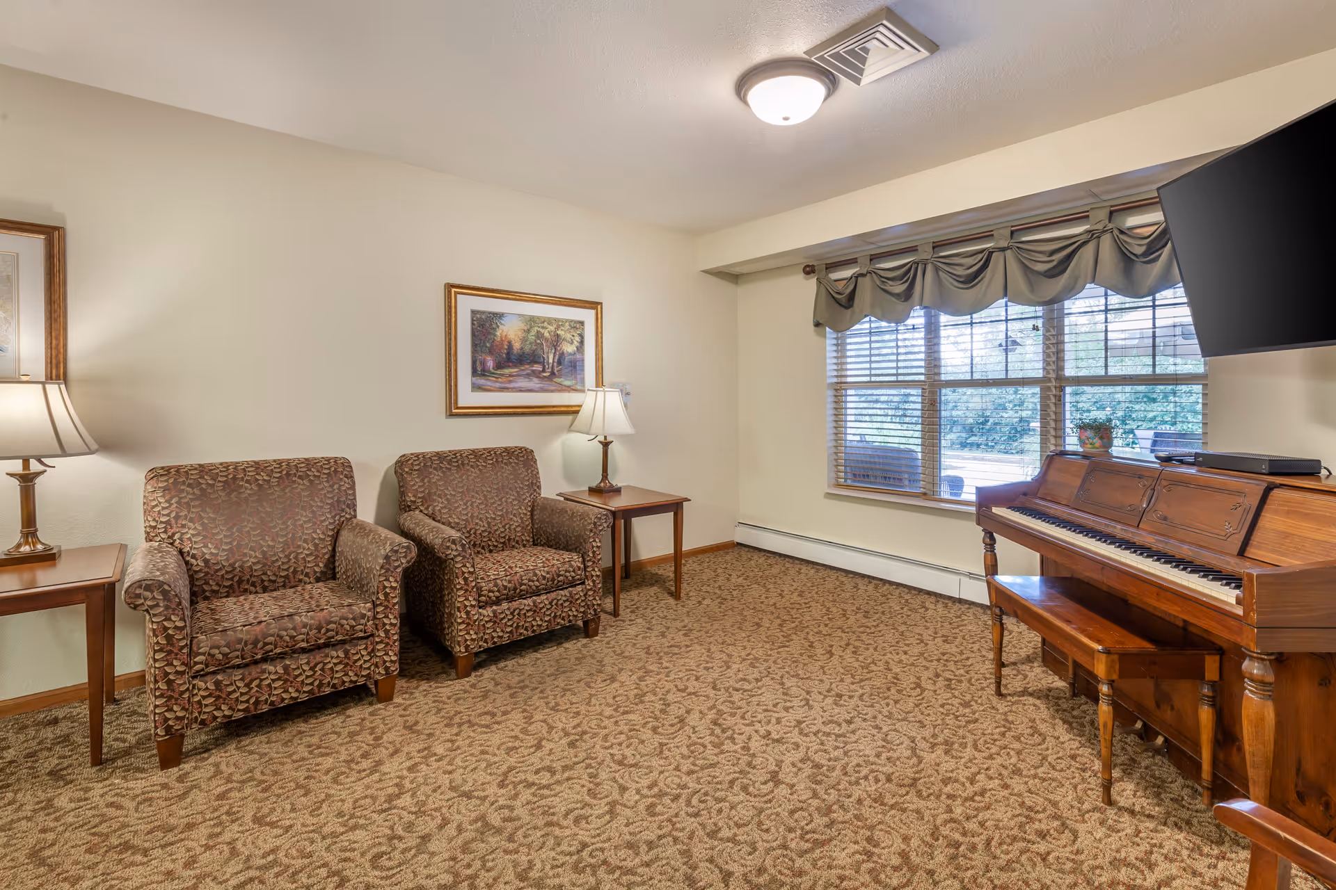 A cozy living room with two patterned armchairs separated by a wooden side table with a lamp. A framed painting hangs on the wall above the chairs. To the right, there is a wooden piano with a bench and a flat-screen TV mounted above it. A large window with valance curtains lets in natural light and shows greenery outside.