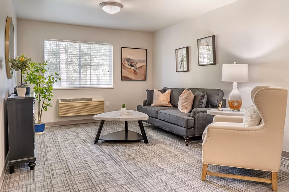 A bright and cozy living room with a gray sofa adorned with pink and gray pillows, a white armchair with a cushion, a round coffee table with a small plant, a side table with a lamp, a window with blinds, framed artwork on the walls, a potted plant, and a patterned carpeted floor.