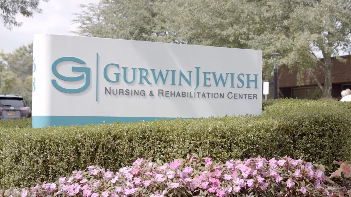 Outdoor view of a large sign for Gurwin Jewish Nursing & Rehabilitation Center surrounded by green bushes and pink flowers, with trees and part of a building in the background.