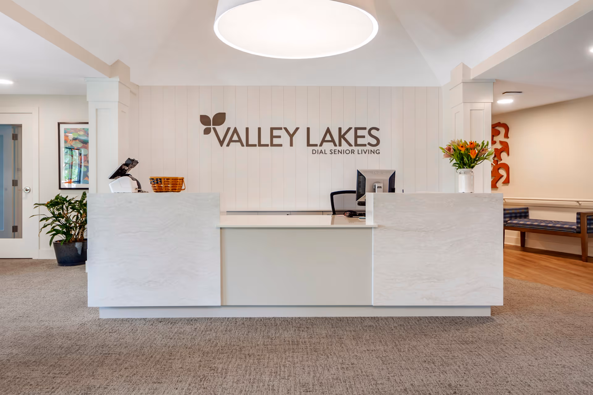 Reception desk area inside Valley Lakes Assisted Living facility with a modern white desk, a computer monitor, a basket, a vase with flowers, and the Valley Lakes Dial Senior Living logo on the wall behind the desk. There is a plant to the left and seating area with a bench to the right.